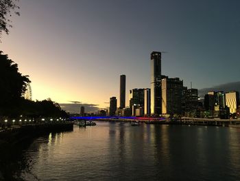 Illuminated buildings by river against sky during sunset