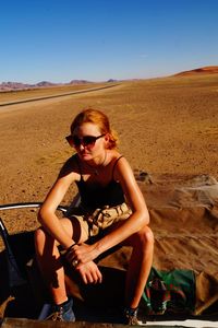 Young woman in sunglasses sitting on desert against clear sky