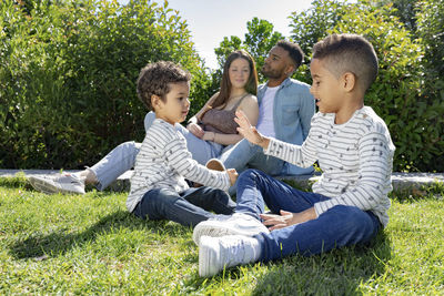 Content ethnic parents with boys looking away and talking while sitting between bushes and lawn in sunlight