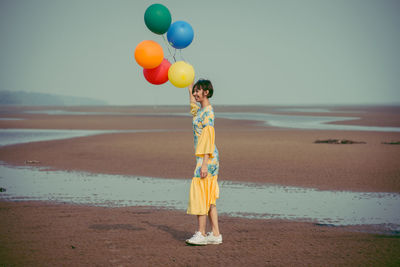 Side view full length of young woman holding colorful balloons at beach