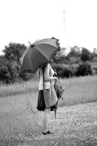 Side view of mature woman with umbrella standing on grassy field against sky
