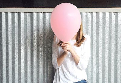 Woman covering face with pink balloon against fence