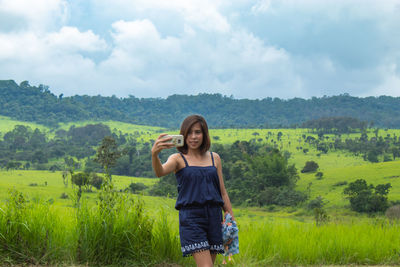 Woman taking selfie while standing on green landscape against cloudy sky