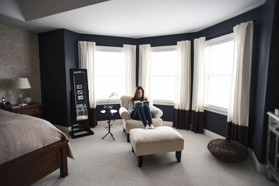 Woman sitting in chair reading book in front of windows in a bedroom.