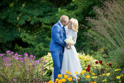 Bridal couple kissing while standing amidst flowers blooming at park