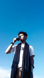 Low angle view of thoughtful young man drinking coffee standing against blue sky