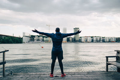 Rear view of male athlete standing with arms outstretched on pier over sea in city