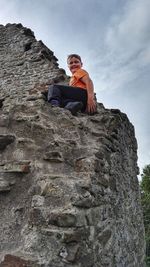 Low angle portrait of boy sitting on rock against sky