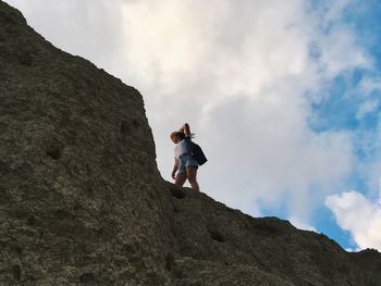 Low angle view of female hiker standing on rocky mountain against cloudy sky