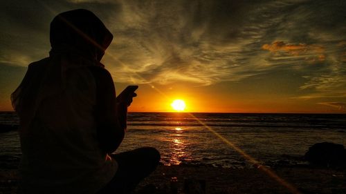 Silhouette man using mobile phone while sitting on beach against sky during sunset