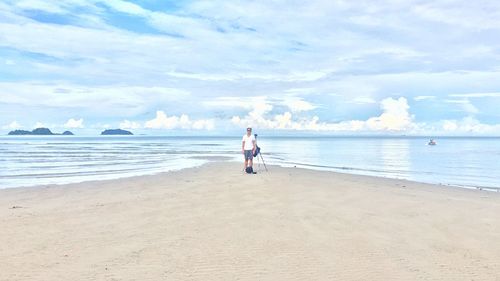 Man standing with tripod at beach against cloudy sky