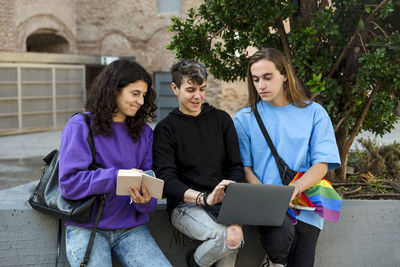 Young diverse people with lgbt rainbow flag using laptop and book outdoors.