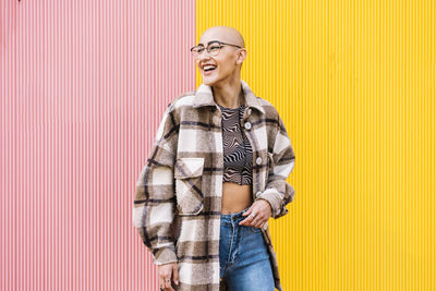 Happy woman with shaved head wearing plaid jacket standing in front of wall