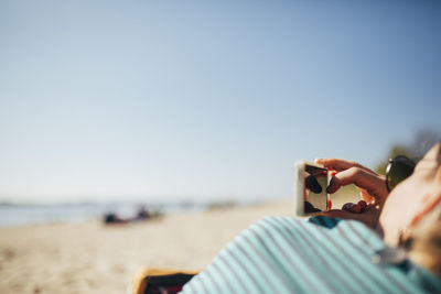 Midsection of woman holding smart phone at beach against clear sky