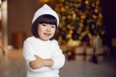 Beautiful korean child girl in in a white knitted sweater and hat stands at the christmas tree 