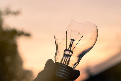 Close-up of hand holding broken light bulb against sky during sunset