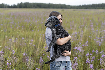 Young smiling woman holding fluffy senior dog in hands on meadow with flowers of fireweed , walking 
