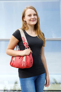 Portrait of smiling woman with shoulder bag standing against glass