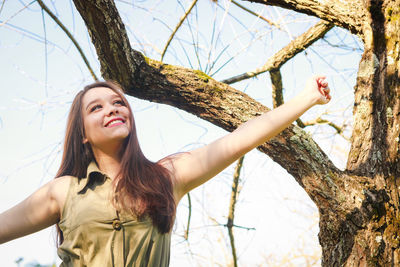 Smiling woman with arms outstretched standing against bare tree