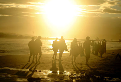 Silhouette people walking at beach against bright sun in sky during sunset