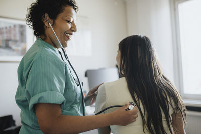 Side view of smiling mature female doctor examining young woman with stethoscope in clinic