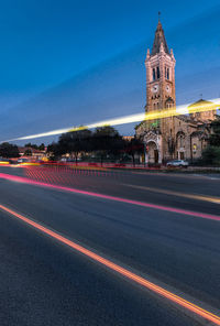 Light trails on road by building against blue sky