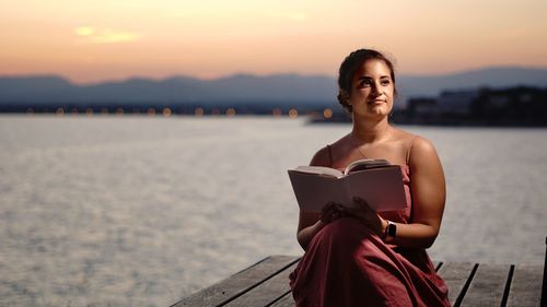 Portrait of smiling woman sitting against sky during sunset reading a book