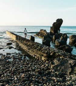 Young man walking on rocks at beach against sky during sunset