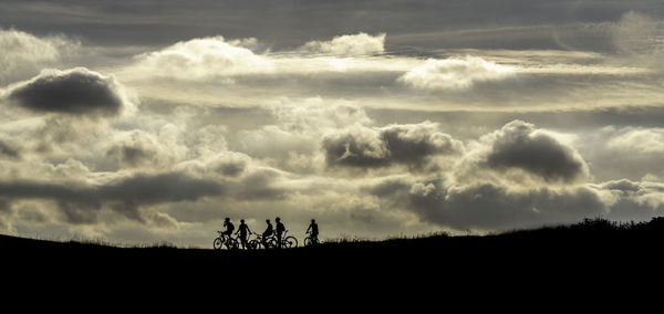 Silhouette people riding bicycles on field against cloudy sky during sunset