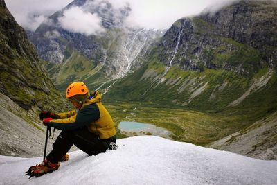 Full length of hiker sitting on glacier against rocky mountains