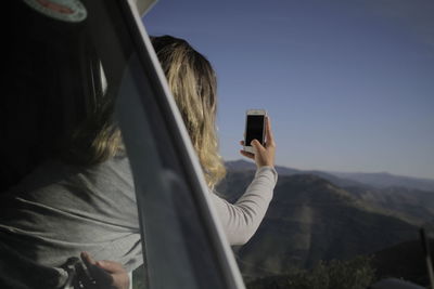 Side view of woman photographing through mobile phone against sky