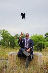 Full length of student throwing mortarboard while sitting on structure at field