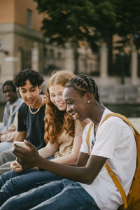 Happy teenage boy with braided hair showing smart phone to female and male friend