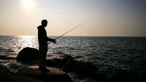 Silhouette man fishing at beach against sky during sunset