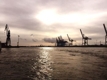 Silhouette cranes at harbor against sky during sunset
