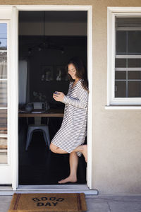 Happy woman holding coffee cup while standing at doorway