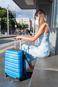 Side view of woman using mobile phone while sitting with suitcase by road