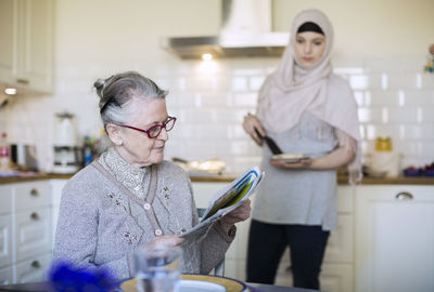Senior woman reading newspaper with female home caregiver preparing food in kitchen