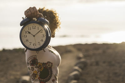 Woman holding clock while standing on field against sky