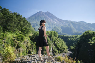 Full length of woman looking away while standing on rock against mountains and sky