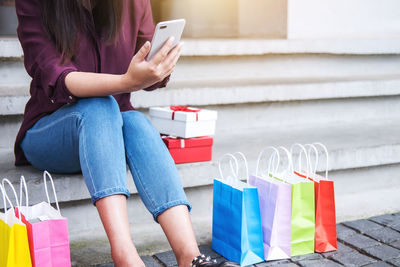 Midsection of woman using smart phone while sitting by colorful shopping bags on steps