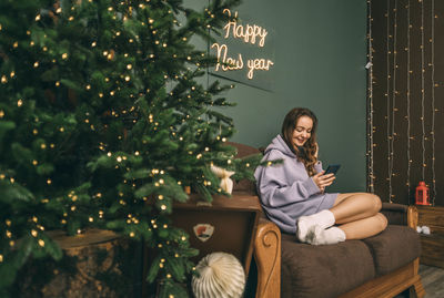 Young woman in headphones listens music on sofa smiling and using phone, near glowing christmas tree