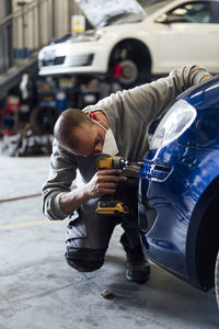 Auto mechanic repairing car with electric screwdriver in workshop