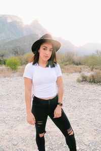Portrait of young woman wearing hat while standing on field against mountains