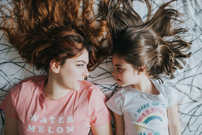 From above young elder sister and funny kid in pajamas looking at each other and lying on bed in bedroom