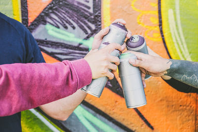 Cropped hands of friends holding spray paint bottles against graffiti wall