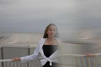 Portrait of young woman standing on railing against sky