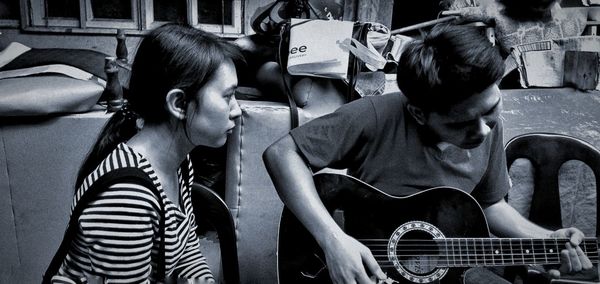 Woman looking at man playing guitar while sitting on chairs