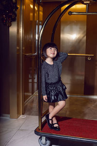 Beautiful korean girl child in a black dress is sitting on a luggage trolley in a hotel