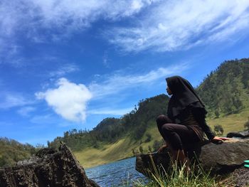 Woman sitting on rock looking at mountain against sky